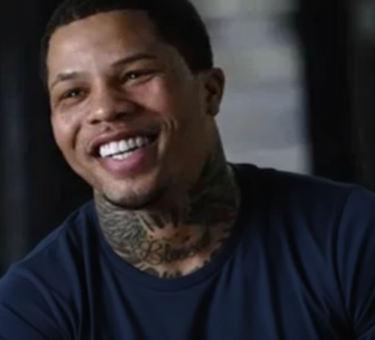 Smiling professional boxer with neck tattoos wearing a dark blue shirt, captured during an interview in an indoor setting.